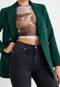 Personne portant un blazer vert foncé, un haut court abstrait marron et beige, et un jean noir taille haute, main posée sur la hanche.