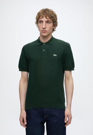 Jeune homme aux cheveux blonds bouclés portant un polo vert foncé à manches courtes et un jean bleu foncé, debout devant un fond blanc uni.