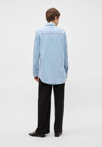 Ανοιχτό μπλε τζιν πουκάμισο με αιχμηρό γιακά, καμπυλωτό στρίφωμα, κουμπωτές μανσέτες και ραφή στην πλάτη, συνδυασμένο με μαύρο παντελόνι wide-leg και επίπεδες παπούτσια.