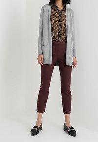Kvinna i grå kardiagan, leopardmönstrad blus, burgundy avkortade byxor och svarta loafers med silversnodder, som står framför en enfärgad bakgrund.