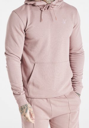 Mann trägt hellrosa Hoodie und passende Sweatpants, mit Händen in den Taschen und sichtbaren Tätowierungen an rechter Hand und am Hals.