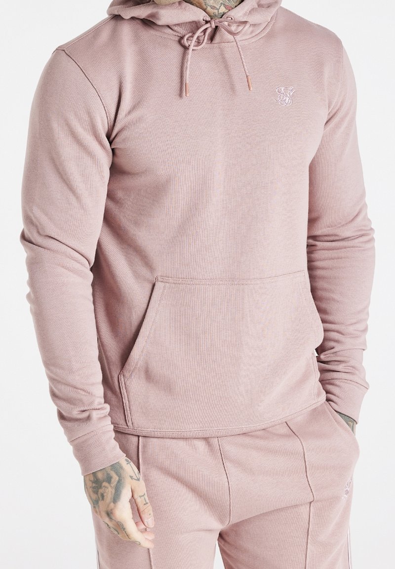 Homme portant un sweat à capuche rose clair et un pantalon de survêtement assorti, les mains dans les poches, avec des tatouages visibles sur la main droite et le cou.