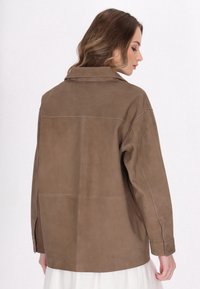 Bruine suède jas met een relaxte pasvorm, puntige kraag en zichtbare naaddetails. Lange mouwen met een knoopmanchet en geen zichtbare hardware.