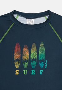 Námornícky modré surf tričko so zelenými akcentmi, na ktorom sú zobrazené štyri farebné surfové dosky s paliem a text "SURF" v živých farbách.
