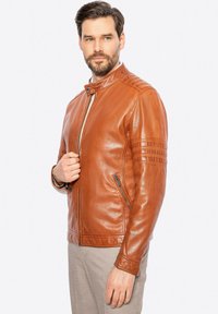 Giacca di pelle marrone con colletto alto, chiusura a zip e tasche laterali. Presenta dettagli cuciti sulle spalle e sulle maniche. Tessuto liscio.