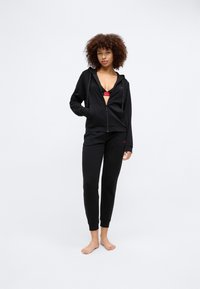 Svart zip-up hoodie med fickor och röd logga, matchad med svarta joggers. Materialet ser mjukt ut; modellen står barfota på en enkel bakgrund.