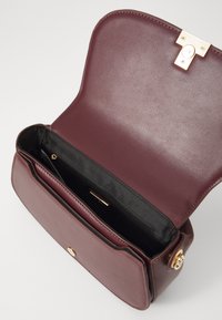 Borsa a mano in pelle bordeaux con una texture liscia, hardware dorato e un unico scomparto interno con zip. La chiusura a pattina presenta cuciture minime.