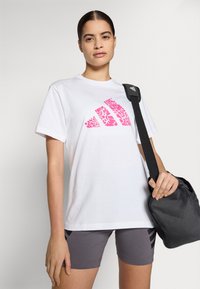 Bílá bavlněná tričko s růžovým květinovým logem Adidas, spárované se šedými šortkami s černými pruhy. Model drží černou tašku.
