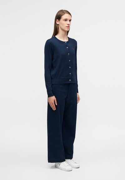 Μπλε ναυτικό ζακέτα με στρογγυλό λαιμόκοψη, κουμπιά μπροστά και μικρό λογότυπο. Συνδυασμένη με φαρδιά μπλε ναυτικά παντελόνια και λευκά αθλητικά παπούτσια.