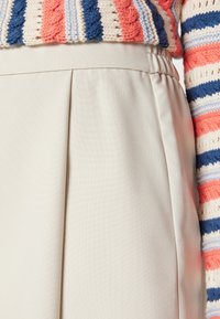 Jupe beige plissée avec une taille élastique, affichant une texture lisse. Le haut en maille présente des rayures horizontales multicolores.