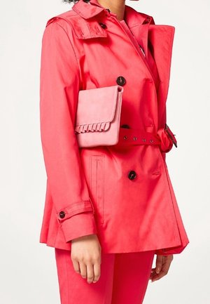 Femme portant un trench-coat ceinturé rose vif avec des boutons noirs, tenant une petite pochette rose sous le bras.