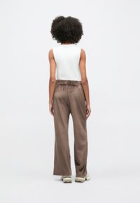 Άτομο με σγουρά μαλλιά που φοράει λευκό αμάνικο τοπ, άνετο καφέ παντελόνι και αθλητικά παπούτσια, στέκεται πλάτη σε ένα απλό φωτεινό φόντο.