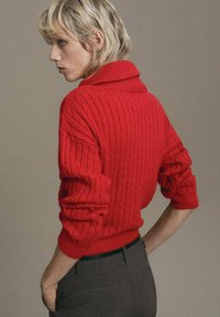 Suéter de punto trenzado rojo con un cuello alto y mangas enrolladas, combinado con pantalones grises. Patrón texturizado en toda la prenda.