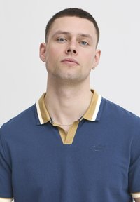 Mornarsko polo majica z kontrastnim zlatim in belim črtastim ovratnikom, ki se ponaša s strukturo tkanine in vezenim logotipom na prsih.