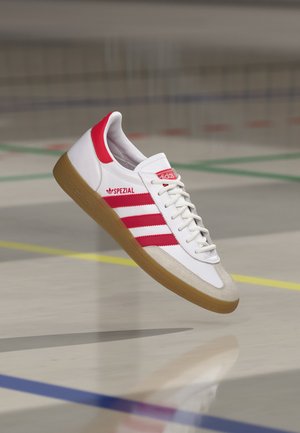 Baskets Adidas Spezial blanches avec des bandes et des accents rouges, embout beige et semelle en gomme, flottant au-dessus d’un sol de gym avec des lignes de terrain colorées.