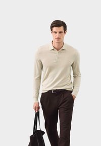 Lichtbeige polo met lange mouwen en drie knopen, gecombineerd met donkerbruine broek. Model houdt een zwarte schoudertas vast. Glad textuur.