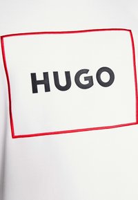 Valkoinen t-paita, jossa on musta "HUGO"-logo punaisessa suorakaiteen muotoisessa kehyksessä. Pehmeä kangas, jolla on sileä pinta ja kontrastiset värit.