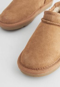Scarpe slip-on in suede marrone con suole in gomma testurizzata, punte arrotondate e dettagli di cucitura lungo i bordi. Fodera interna morbida visibile.