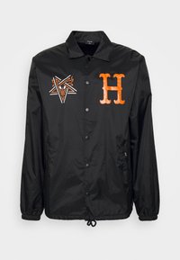 Veste noire à boutons avec "H" stylisé orange sur la poitrine droite et motif étoile orange et blanc sur la poitrine gauche, poignets élastiques et ourlet avec cordon de serrage.