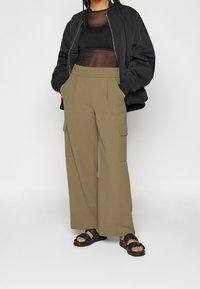 Olive green cargo pants with a wide leg, featuring side pockets. Paired with a black sheer top and a black bomber jacket. Black sandals.