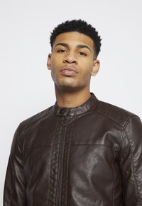 Chaqueta de cuero marrón con cuello camisero, cremallera frontal y acentos de costura horizontal. Textura suave y corte ajustado.