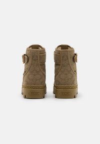 Botte beige avec un dessus en toile texturée, semelle en caoutchouc et accents en métal doré. Elle présente une boucle et un motif sur la surface du matériau.