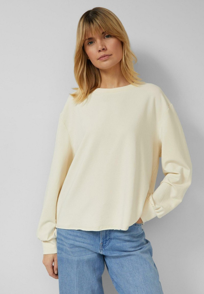 Blondide juustega naine kannab lahtist koorevärvi pika varrukaga särki ja helesiniseid teksapükse, seistes tavalise halli tausta ees.