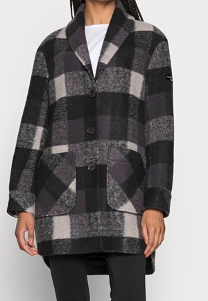 Mannequin portant un long manteau à carreaux noir, gris et crème, à larges revers et trois boutons, sur un haut blanc et un pantalon noir.
