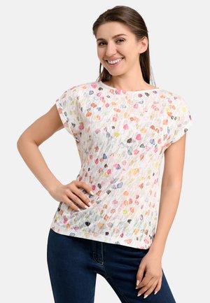 White short-sleeve top with multicolored abstract patterns. Lightweight, textured fabric, crew neckline, and relaxed fit. Paired with dark blue jeans.