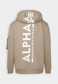 Bež kapuljača s velikim belim logotipom "ALPHA INDUSTRIES" vertikalno odtisnutim na zadnjem delu. Ima kapuljaču s vezicama in žep na zunanji strani rokava.