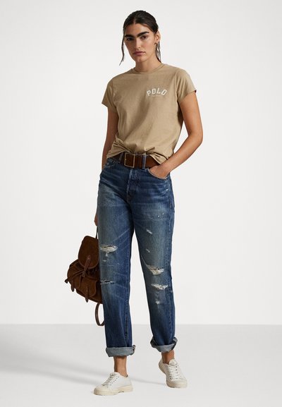 Beżowa bawełniana koszulka z napisem "POLO", zestawiona z przetartymi niebieskimi dżinsami, podwiniętymi mankietami, brązowym paskiem i białymi trampkami. Brązowa torba z zamszu.