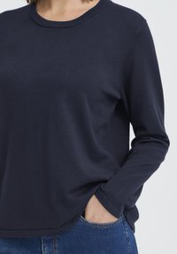 Pull à manches longues bleu marine en tissu maille douce, doté d'un col rond, d'une coupe ample et de fentes sur les côtés, porté avec un jean.