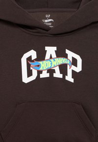 Καφέ φούτερ με κουκούλα με λευκό λογότυπο «GAP» και πολύχρωμο γραφικό «Hot Wheels» στο στήθος· διαθέτει τσέπη μπροστά και απαλής υφής ύφασμα.
