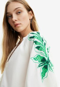 Blusa bianca con intricate ricami a foglia verde sulle maniche e dettagli in perline. Tessuto leggero, vestibilità ampia e scollatura a V.