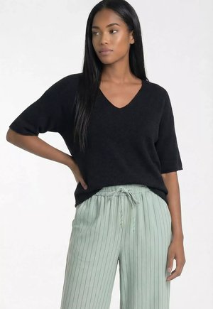 Kobieta o długich czarnych włosach, ubrana w czarny top z krótkim rękawem i dekoltem w serek oraz jasnzie zielone spodnie w drobne prążki ze sznurkiem, stoi z ręką na biodrze.