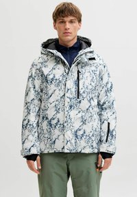 Veste isolante à capuche à motifs blancs et bleus, avec fermeture éclair et plusieurs poches, portée par-dessus une couche de base sombre et un pantalon vert.