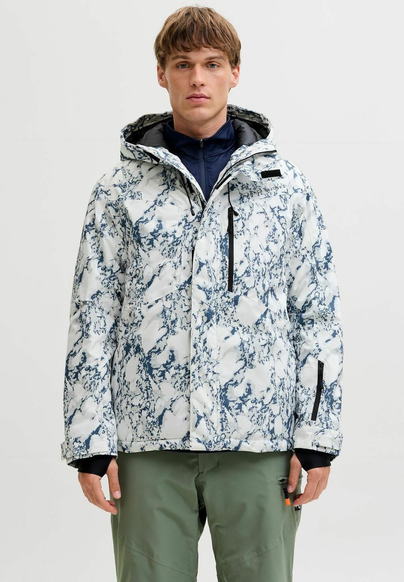 Veste isolante à capuche à motifs blancs et bleus, avec fermeture éclair et plusieurs poches, portée par-dessus une couche de base sombre et un pantalon vert.