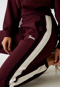Bordeaux sportbroek met witte zijkanten en zwarte accenten; beschikt over een elastische tailleband en geborduurd "Guess" logo.