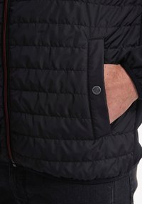 Giacca imbottita nera con zip frontale, dotata di tasca frontale e accento con logo rotondo. Il tessuto ha una texture liscia con cuciture orizzontali.