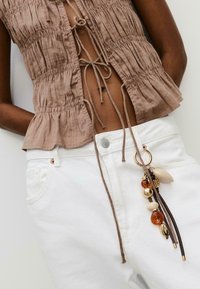 Haut smocké marron avec un devant noué, associé à un denim blanc. Accessoire de porte-clés avec des perles, des accents en cuir et un charme en or.