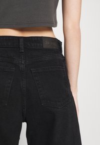 Mustat farkut korkealla vyötäröllä, joissa on MONKI-logopatchi, takataskut ja hieman haalistu kankaan pinta.