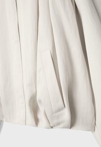 Gros plan d'un tissu beige avec une poche passepoilée inclinée et des détails de couture visibles sur un vêtement.
