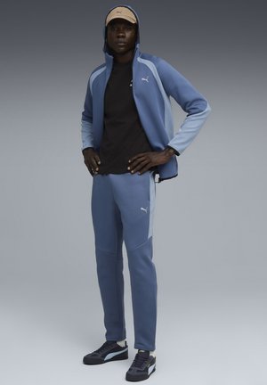 Lichtblauwe trainingspak met een ritsjack en tapered broek. Zwarte shirt eronder, gedragen met een beige pet en zwarte sneakers. Accenten met Puma-logo's.