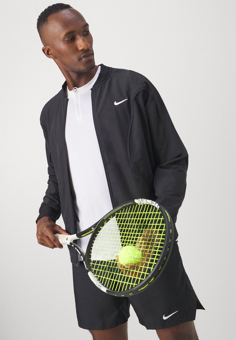 Man in zwarte Nike-jack en shorts met een tennisracket en bal, kijkend opzij tegen een effen achtergrond.