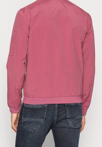 Osoba v růžovém bomberu a tmavě modrých džínách, zobrazená zezadu na jednoduchém bílém pozadí.