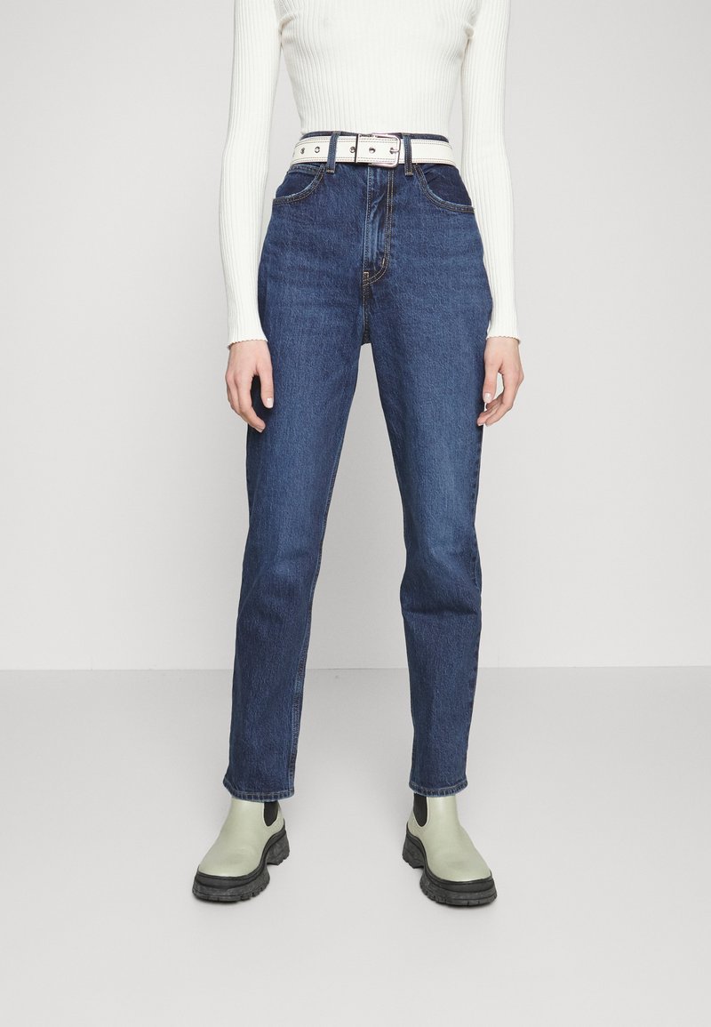 Personne portant un jean bleu taille haute, un haut blanc à manches longues en côte, une ceinture blanche et des bottines vert clair, debout sur un fond uni.