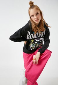 Czarna bluza z kapturem z graficznym nadrukiem przedstawiającym smoka, w zestawieniu z jasnoróżowymi, szerokimi spodniami. Białe, masywne sneakersy i kolorowe pierścionki.