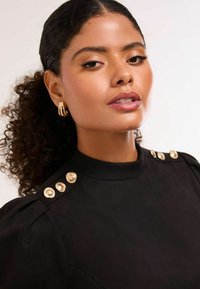 Svart topp med hög krage som har guldknappar på axlarna och texturerad tyg, med en modell som visar upp lockigt hår och hoopörhängen.