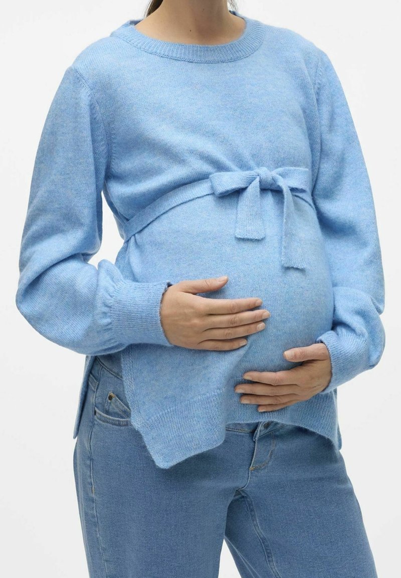 Personne enceinte portant un pull bleu clair et un jean, tenant doucement son ventre avec les deux mains par-dessus et par-dessous.