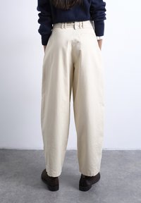 Άτομο που φοράει μπεζ παντελόνι ψηλόμεσο με φαρδιά μπατζάκια, σκούρα καφέ παπούτσια και μπλε πουλόβερ, στέκεται σε γκρι τσιμεντένιο πάτωμα μπροστά σε λευκό τοίχο.